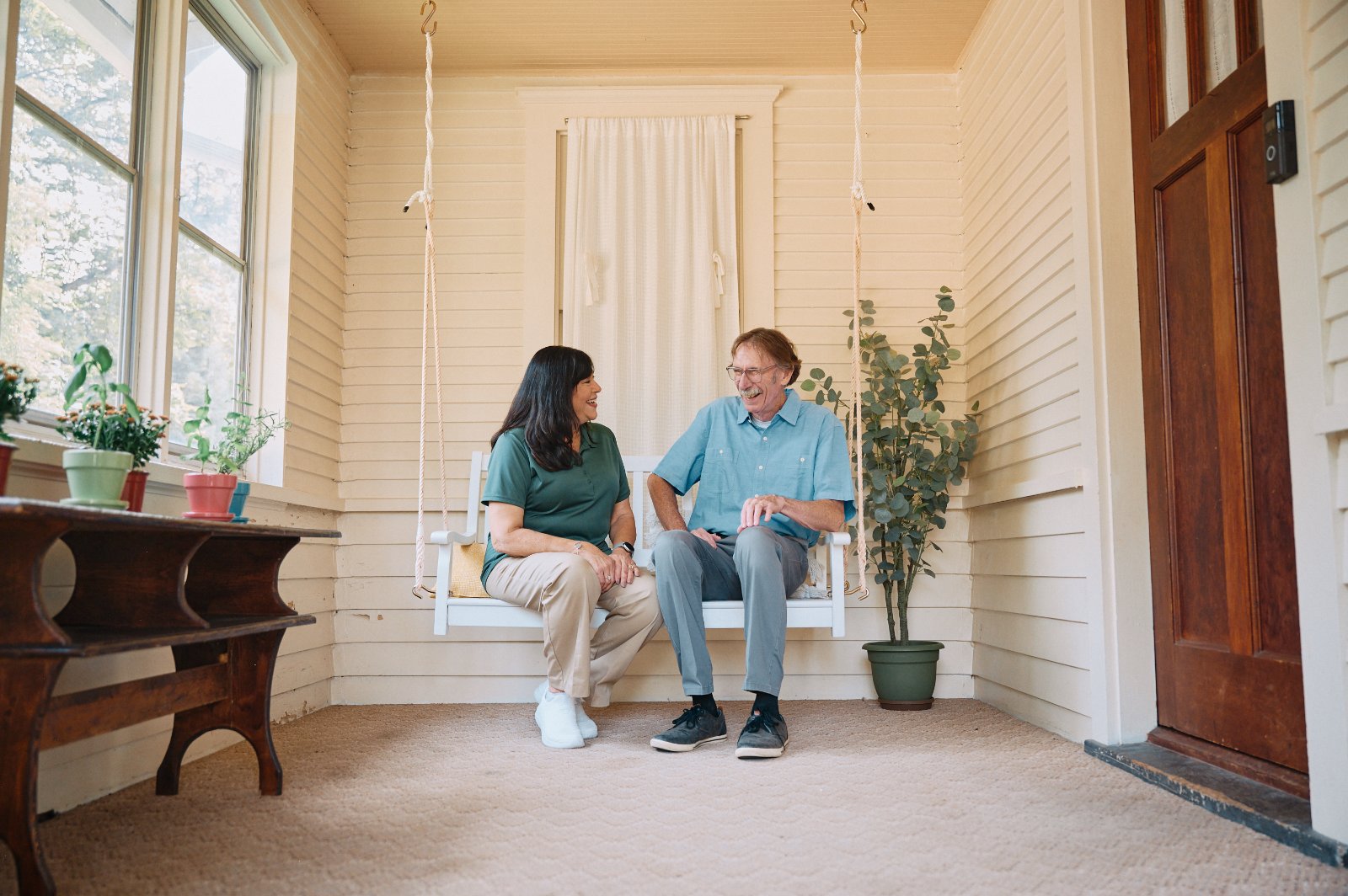 A cheerful elderly woman wearing a sun hat gardens alongside her Home Instead caregiver in a colorful and vibrant outdoor setting.