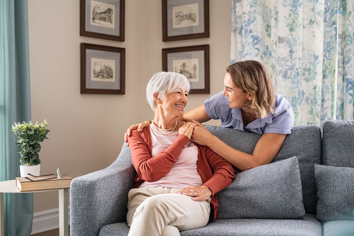elderly woman sitting on couch with a younger woman embracing her from behind