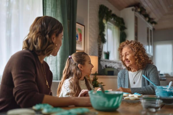 Grandmother, daughter, and granddaughter sit together at a kitchen table, smiling and talking.