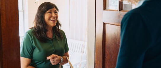 A Home Instead caregiver greets an elderly woman at the door with a warm smile, holding a tote bag, symbolizing friendliness and readiness to assist.