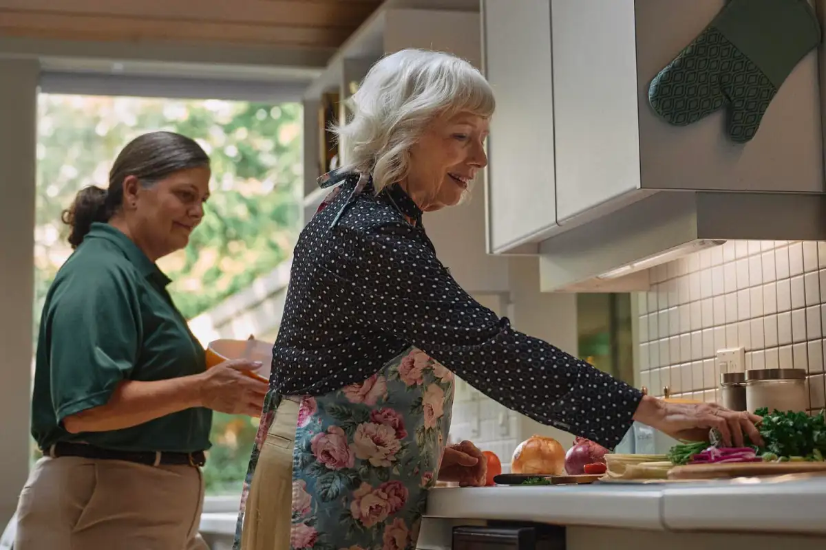 A Home Instead caregiver and an elderly woman prepare a meal together in a sunny kitchen, symbolizing teamwork and independence in home care.