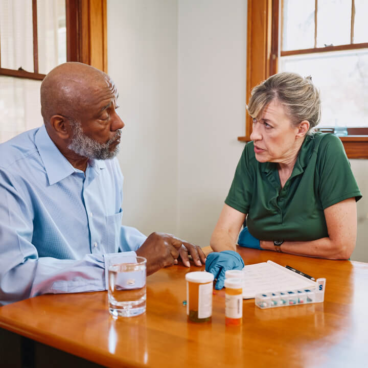 A Home Instead caregiver and an elderly man sit at a table discussing medications, with pill organizers and water, showcasing careful health management.