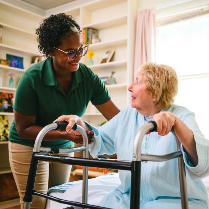 A smiling Home Instead caregiver in a green uniform assists an elderly woman using a walker in a bright and welcoming room, emphasizing support and encouragement.