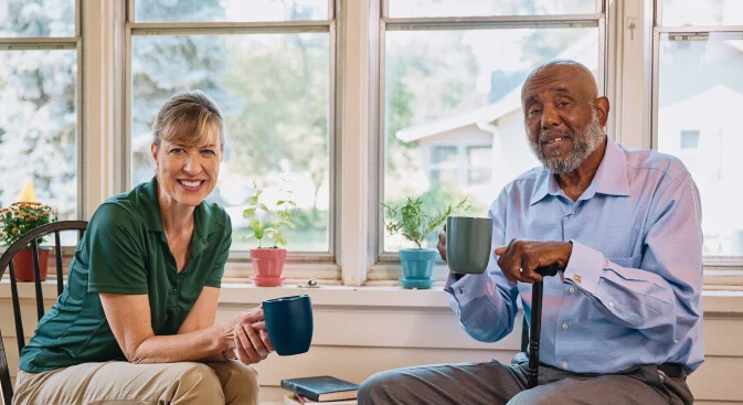 A compassionate caregiver and elderly man smile as they sit and share coffee.