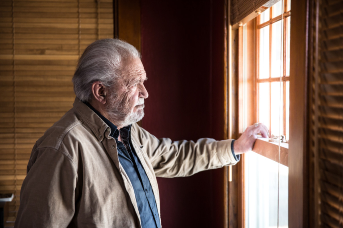 Elderly man gazes out the window of his home.