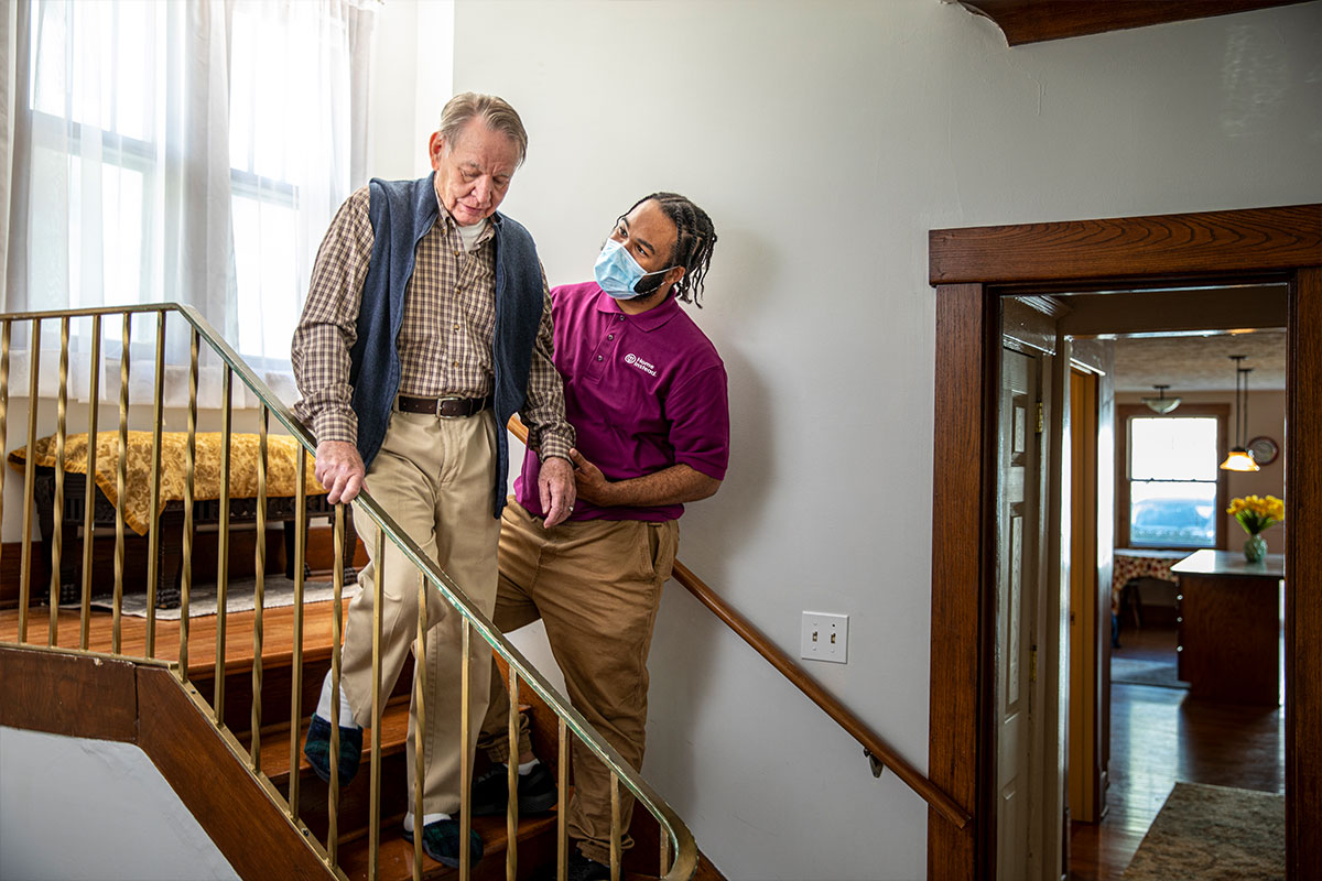 Home Instead Caregiver wearing mask helps senior man walking down stairs at home