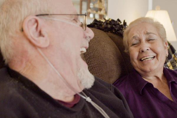 HISC Caregiver and senior man laugh while sitting on couch at home.