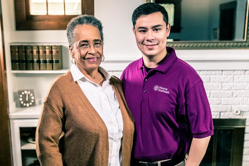 Home Instead Caregiver and senior woman with cane stand smiling together at home