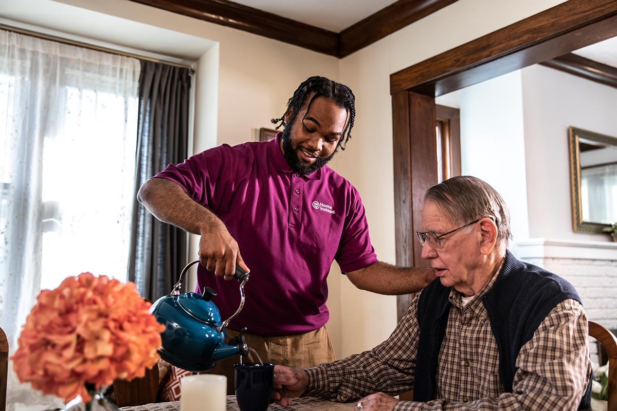 Home Instead Caregiver pours coffee for senior man sitting at home