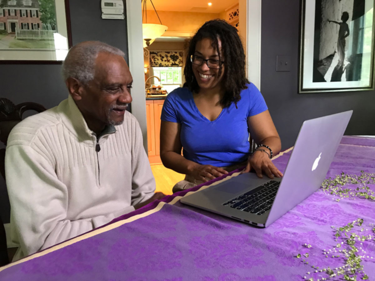 Caregiver and aging man read from laptop on dining room table.